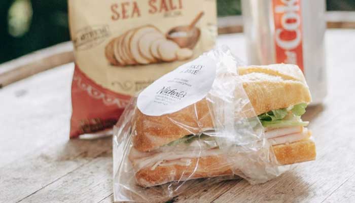 A catering-style sandwich in plastic wrap, chips, and a Diet Coke can on a wooden surface.